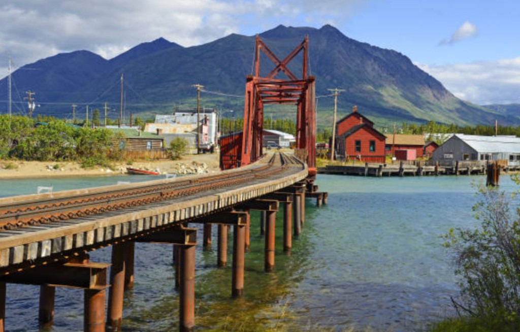 White Pass and Yukon Route railway trestle bridge over turquoise water with historic red buildings and mountain peaks near Carcross, Yukon